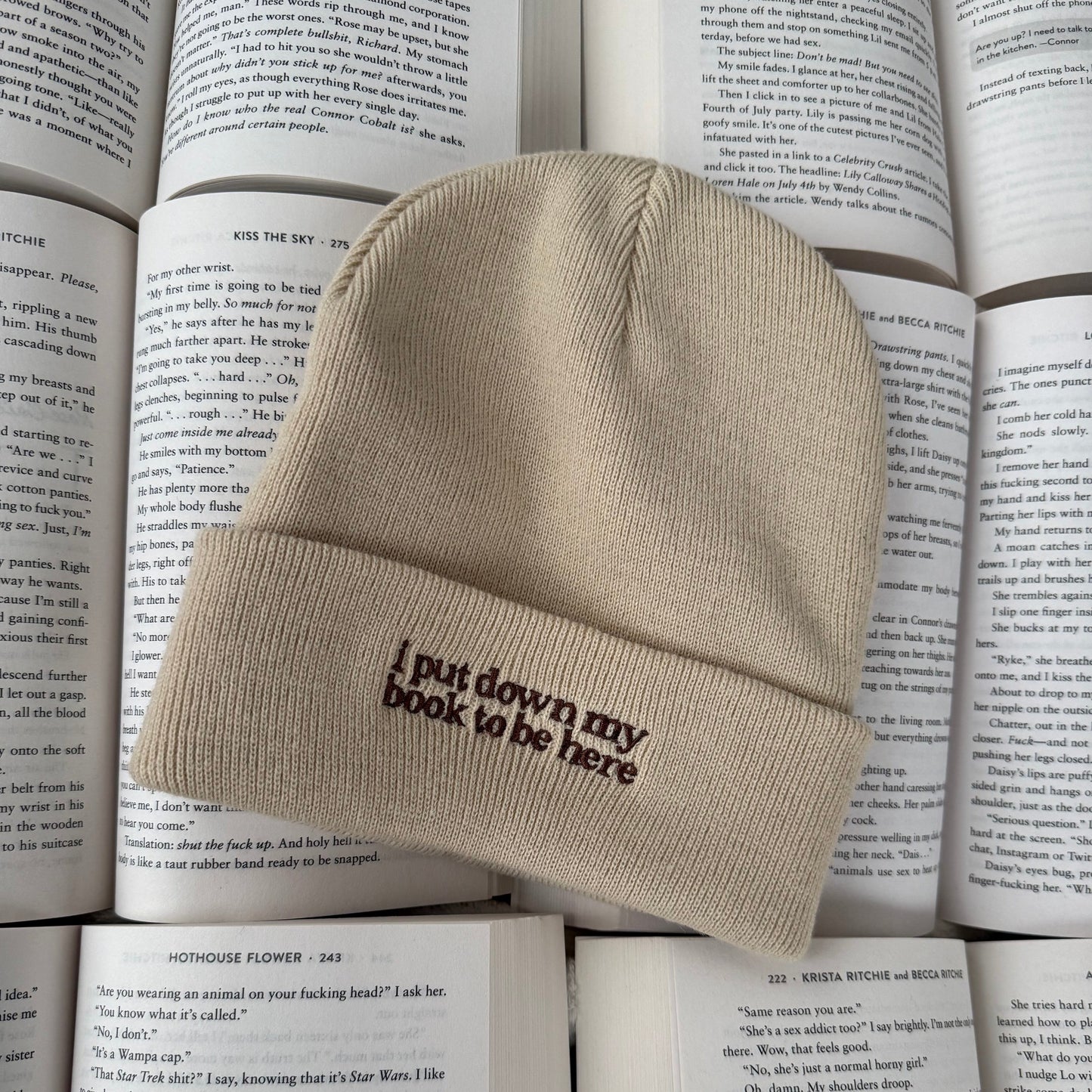 Beige beanie with embroidered text reading "i put my book down to be here" on a stack of books