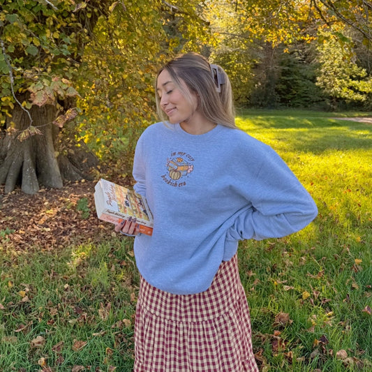 Woman in an embroidered grey sweater and plaid skirt standing in a park with trees and sunlight filtering through.
