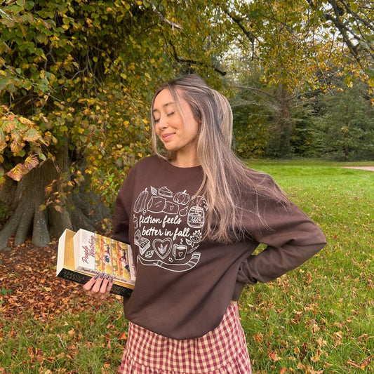 Woman in a park holding a book, wearing a brown sweatshirt with text and a checkered skirt.