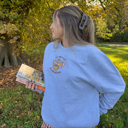 Person wearing a grey sweatshirt with an embroidered design, holding a book, standing in a park with trees and grass.