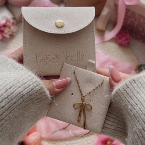 Gold necklace in a 'Page & Needle' branded box held by a person wearing a beige sweater with pink flowers in the background.