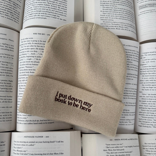 Beige beanie with embroidered text reading "i put my book down to be here" on a stack of books