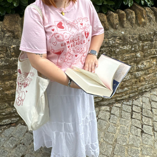 Person wearing a pink 'Romance Reader' t-shirt holding an open book against a stone wall.