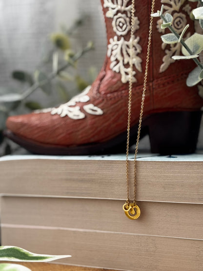 Gold necklace with a horseshoe pendant on a wooden surface next to a brown boot with white lace.