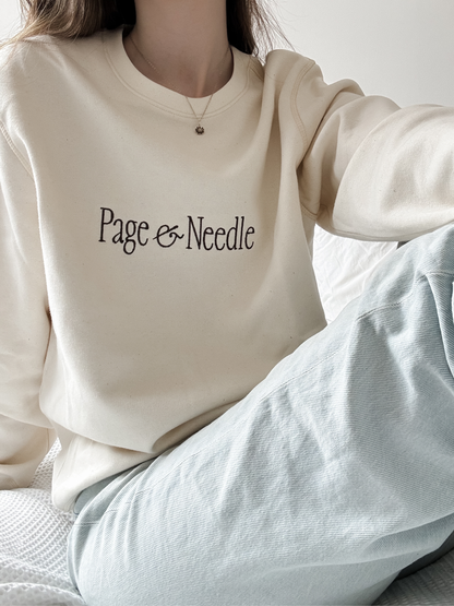 A person wearing a cream-colored crewneck sweatshirt with 'Page & Needle' printed in black script across the chest, paired with light blue textured jeans.