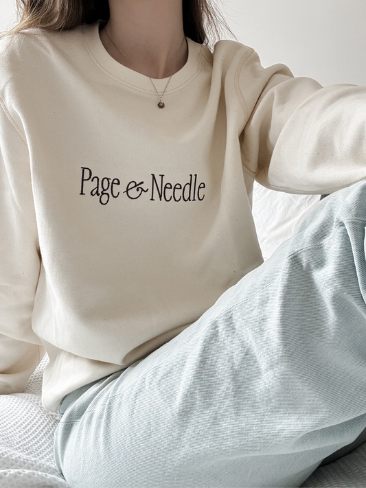 A person wearing a cream-colored crewneck sweatshirt with 'Page & Needle' printed in black script across the chest, paired with light blue textured jeans.