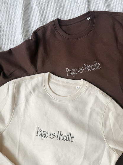 Two crewneck sweatshirts laid flat on a textured white fabric background. The top sweatshirt is dark brown with white script text reading 'Page & Needle', and the bottom sweatshirt is off-white/cream with black script text reading 'Page & Needle'.