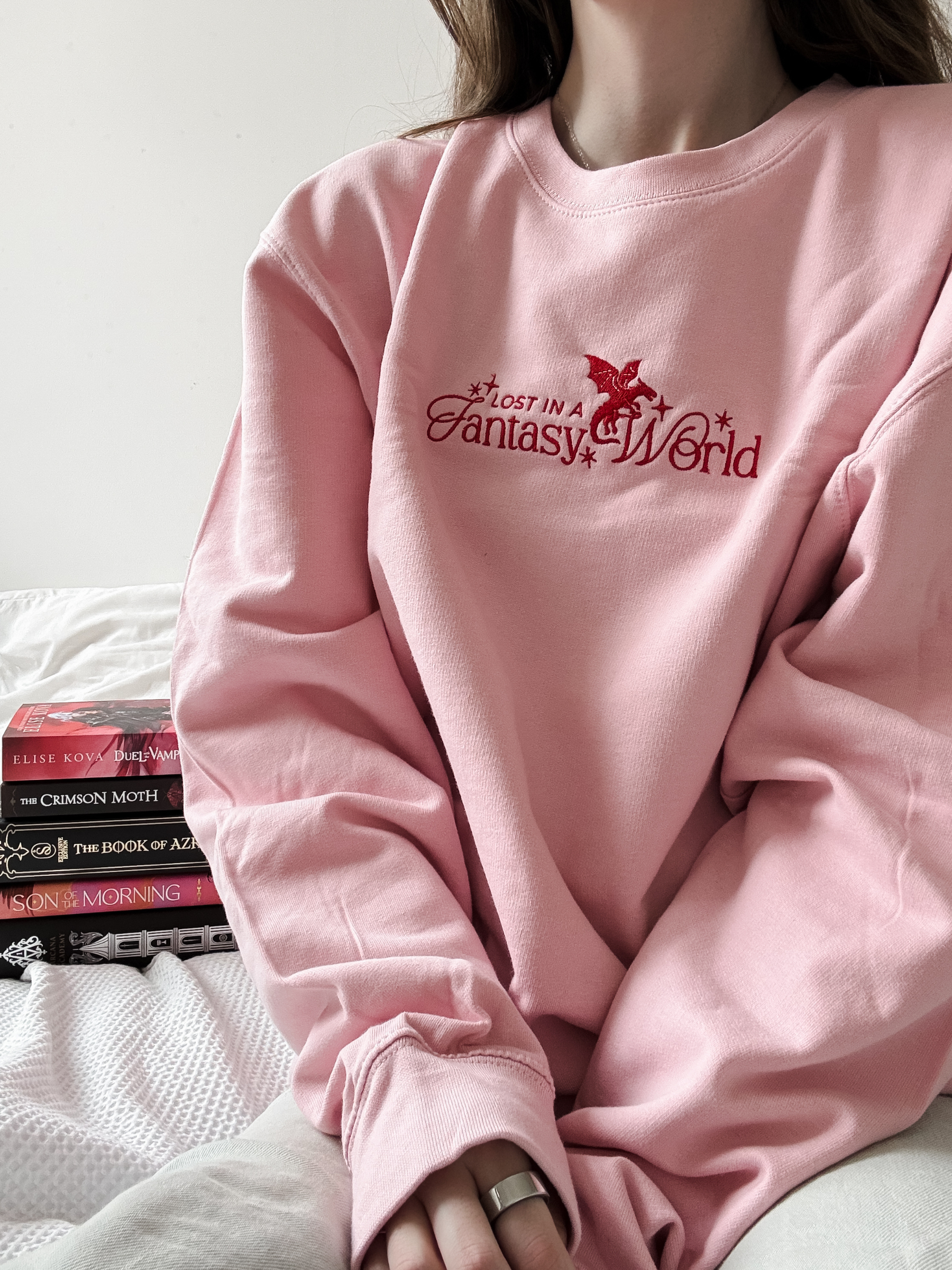 A person wearing an oversized light pink sweatshirt with a red embroidered graphic reading 'Lost in a Fantasy World' on the chest, seated on a bed with books nearby.