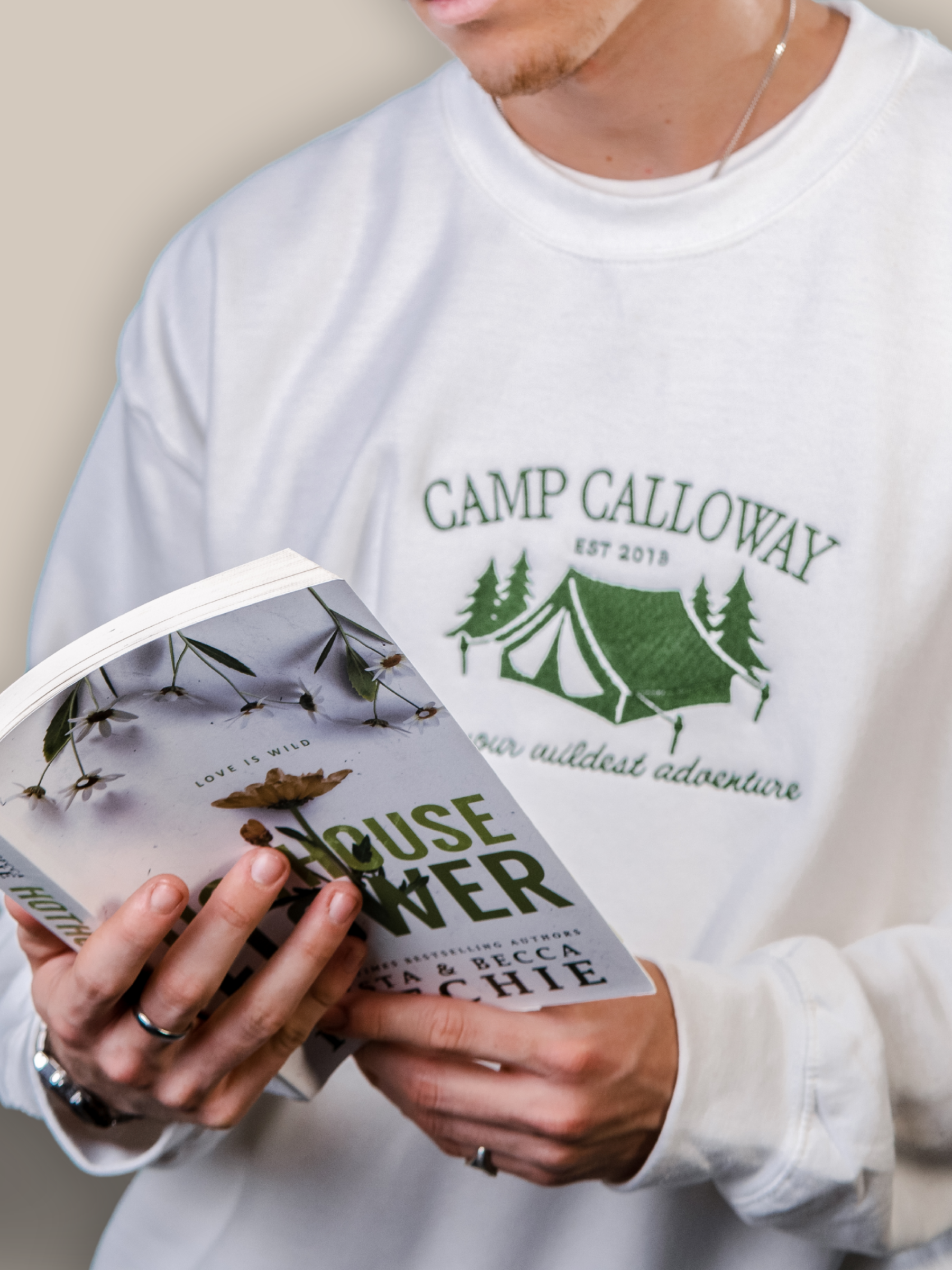 Person wearing a white sweatshirt with 'Camp Calloway' design, holding a book.