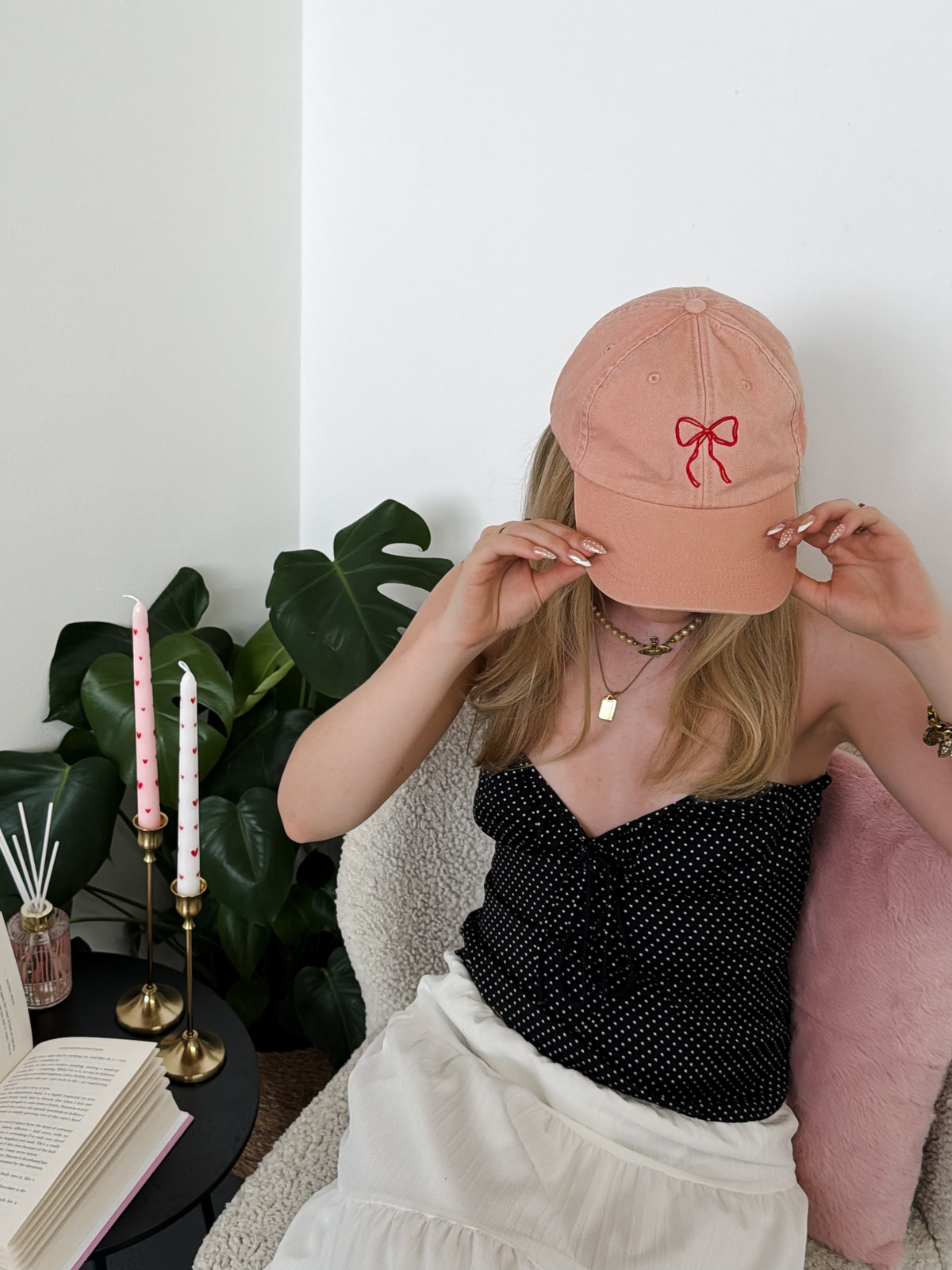 A woman wearing a dusty pink baseball cap with a red embroidered bow, holding it in front of her face while wearing a black polka-dot strapless top and a white skirt in a cozy indoor setting.
