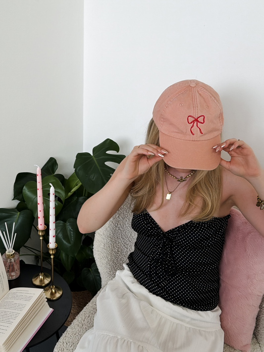 A woman wearing a dusty pink baseball cap with a red embroidered bow, holding it in front of her face while wearing a black polka-dot strapless top and a white skirt in a cozy indoor setting.