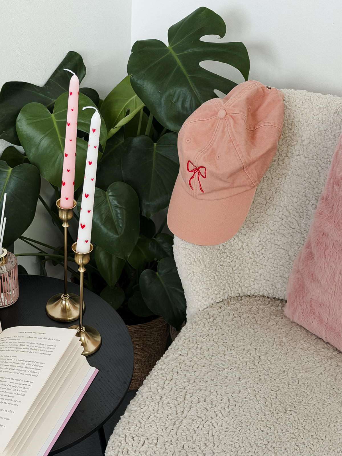 A dusty pink baseball cap with a red bow embroidery sits on a cream sherpa chair next to a pink pillow, with a pink and white candle in a brass holder on a side table in the foreground.